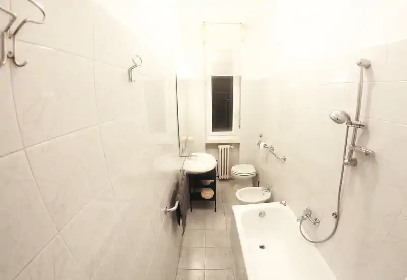 Bright, narrow bathroom with bathtub, wall-mounted shower, sink, bidet and toilet; white tiled walls and a small window at the far end.
