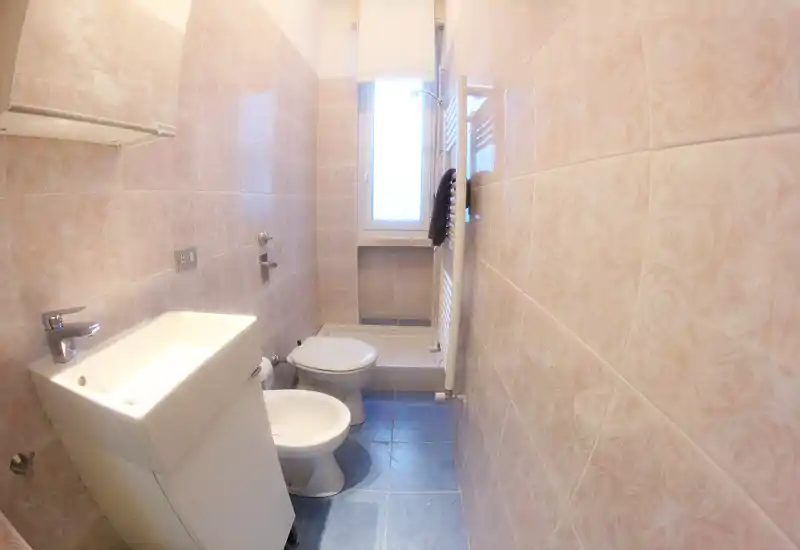 Compact tiled bathroom with sink, bidet, toilet and a small shower under a window; light pink walls and blue floor tiles create a bright, clean look.