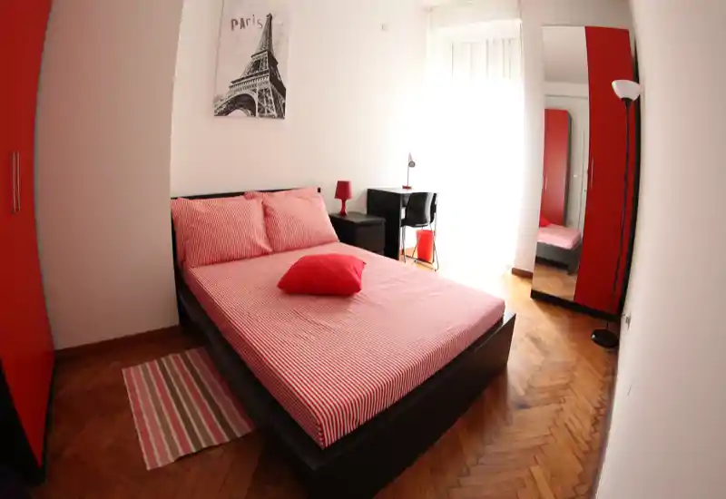 Camera da letto luminosa e arredata con letto matrimoniale con lenzuola rosse a righe, comodino e scrivania vicino a una grande finestra; armadio e specchio a tutta altezza completano l'ambiente.