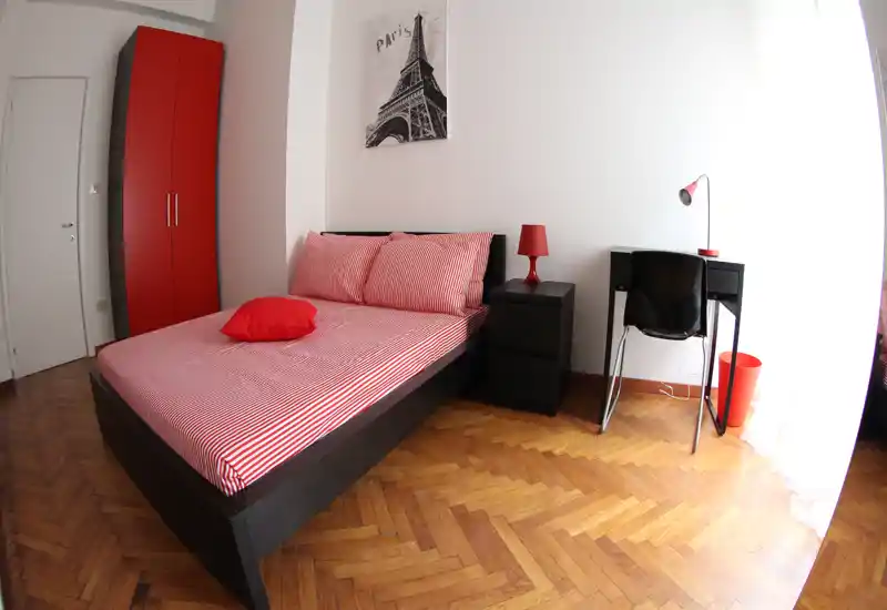 Camera da letto luminosa e moderna con letto matrimoniale con lenzuola a righe rosse, lampada e armadio coordinati; luce naturale abbondante e una scrivania piccola per lavorare.