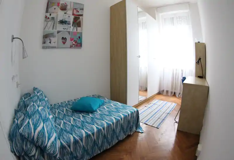 Camera singola compatta e luminosa con copriletto blu a motivi, armadio con specchio e una scrivania vicino alla finestra — ideale per uno studente o un single.
