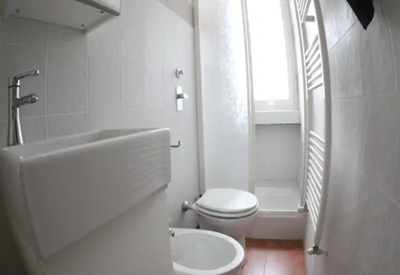 Bagno compatto e luminoso con lavabo, bidet, wc e una piccola doccia vicino alla finestra. Piastrelle bianche e sanitari moderni danno un aspetto pulito.
