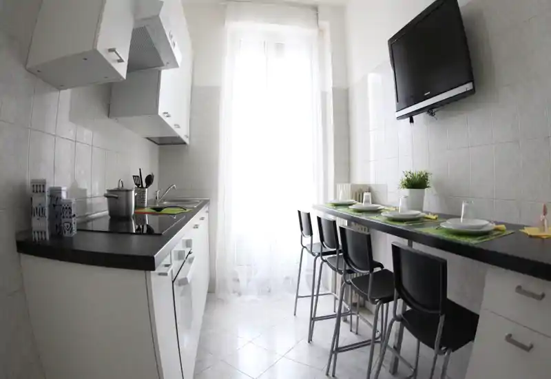 Cucina moderna e compatta con piano nero, piano cottura integrato e un bancone con sgabelli alti. Luce naturale intensa da una finestra alta e arredo minimale rendono lo spazio funzionale e accogliente.