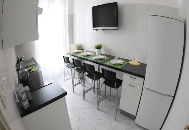 Cucina compatta e moderna con un bancone lungo e quattro sgabelli alti, apparecchiata per la tavola; luce naturale intensa e TV a parete completano l'ambiente.