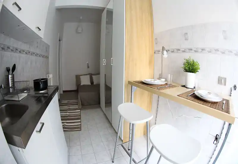 Monolocale compatto con angolo cottura e zona notte. Si vede un letto singolo sullo sfondo, un armadio con specchio e un bancone con sgabelli in primo piano — disposizone pratica per soggiorni brevi.