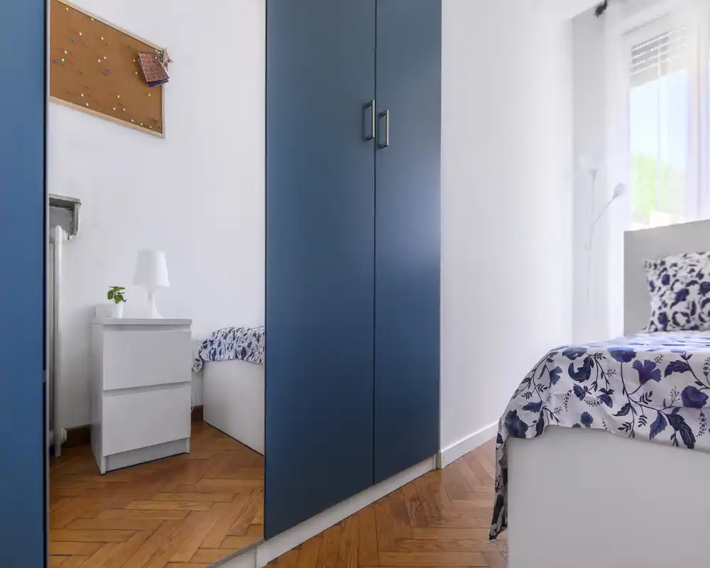 Chambre lumineuse et soignée avec une couette à motif, une table de chevet et une armoire à miroir reflétant une partie de la pièce.