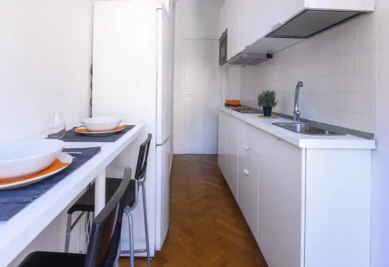 Cucina compatta e moderna con mobili bianchi, lavello integrato e bancone colazione per due; ambiente luminoso e ben organizzato, ideale per monolocali.