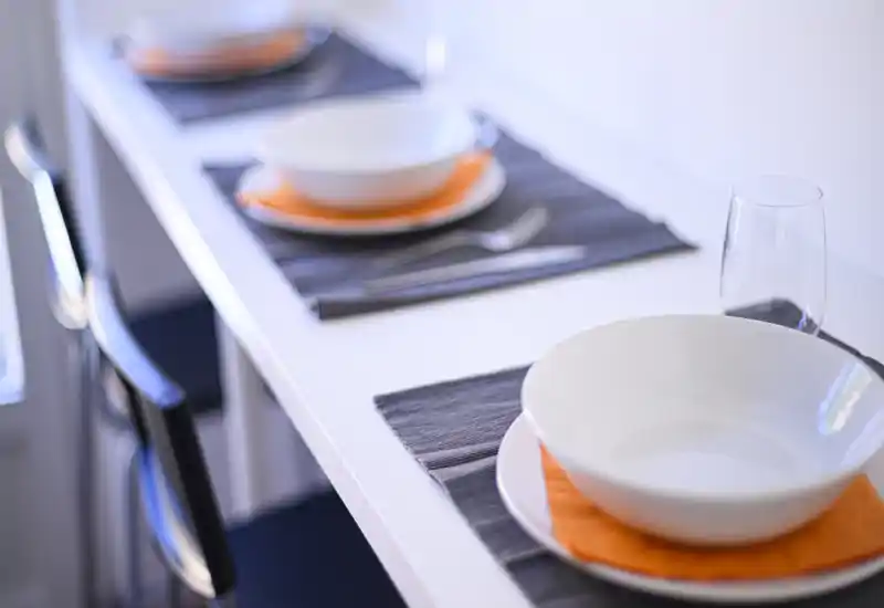 Primo piano di un banco da pranzo moderno apparecchiato: tovagliette, piatti, ciotole e bicchiere su un tavolo bianco stile bancone con sgabelli.