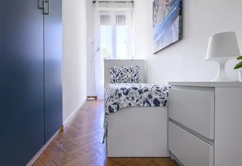 Camera singola luminosa e compatta con letto dal copriletto bianco e blu, mobili bianchi e pavimento in parquet a spina di pesce. La luce naturale dalla finestra rende l'ambiente arioso e ordinato.
