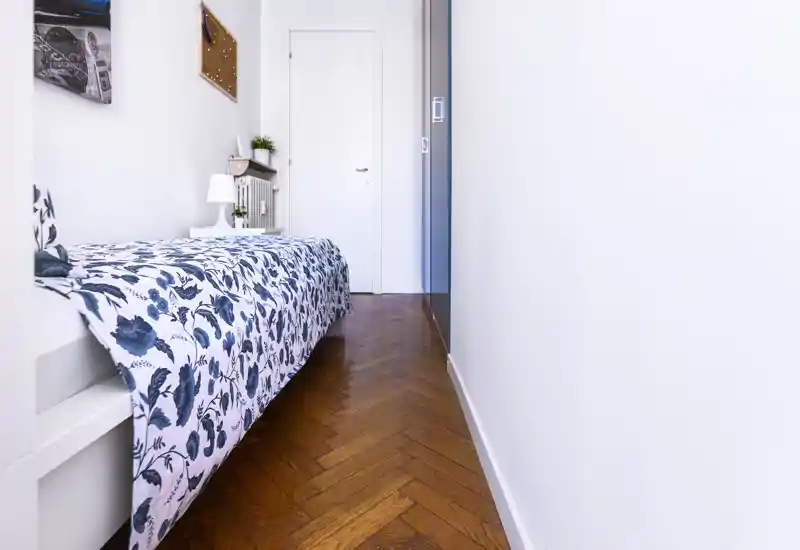 Camera singola compatta e luminosa con copriletto blu e bianco, pavimento a spina di pesce in legno e arredo moderno essenziale.