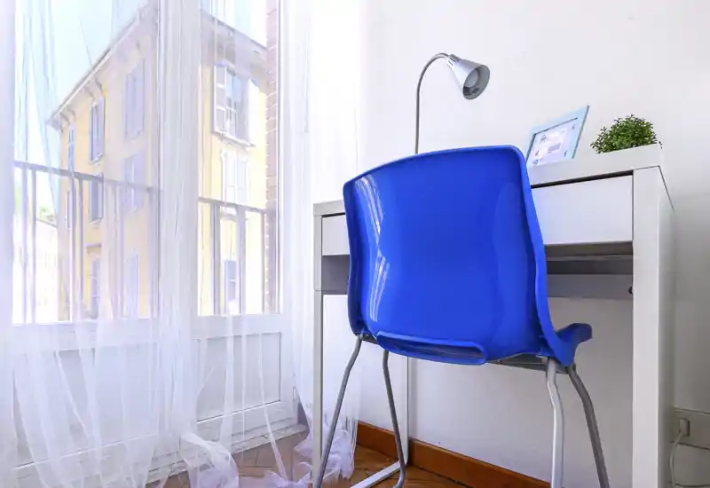 Piccolo angolo studio luminoso vicino a una grande finestra con scrivania bianca, sedia blu, lampada e tende leggere: un angolo accogliente per lavoro o studio.