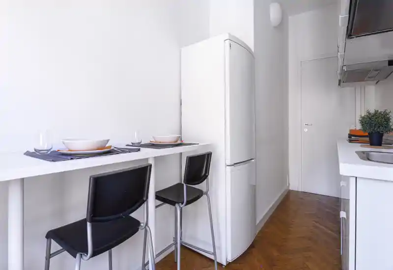 Cucinotto moderno compatto con bancone, due sgabelli, frigorifero e lavello integrato — luminoso, ordinato e ideale per soggiorni brevi.