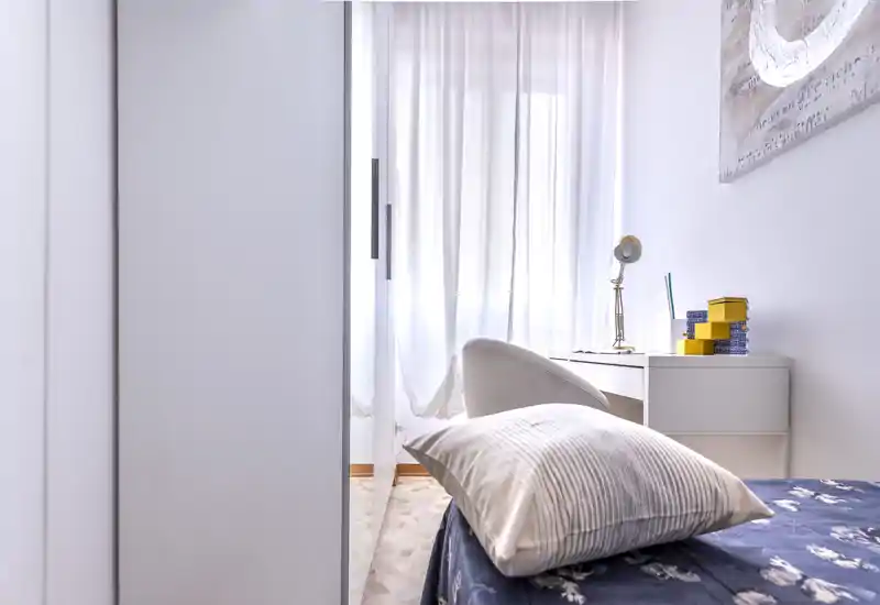 Bright, compact bedroom with a visible single bed, soft pillow and a small desk by a sheer-curtained window providing pleasant natural light.