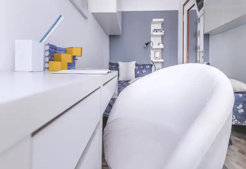 Bright, compact bedroom corner with a white desk and an inviting padded chair facing a small bed/sofa with patterned cushions. The composition highlights a tidy work area and a cozy sleeping nook, ideal for a studio or small apartment listing.