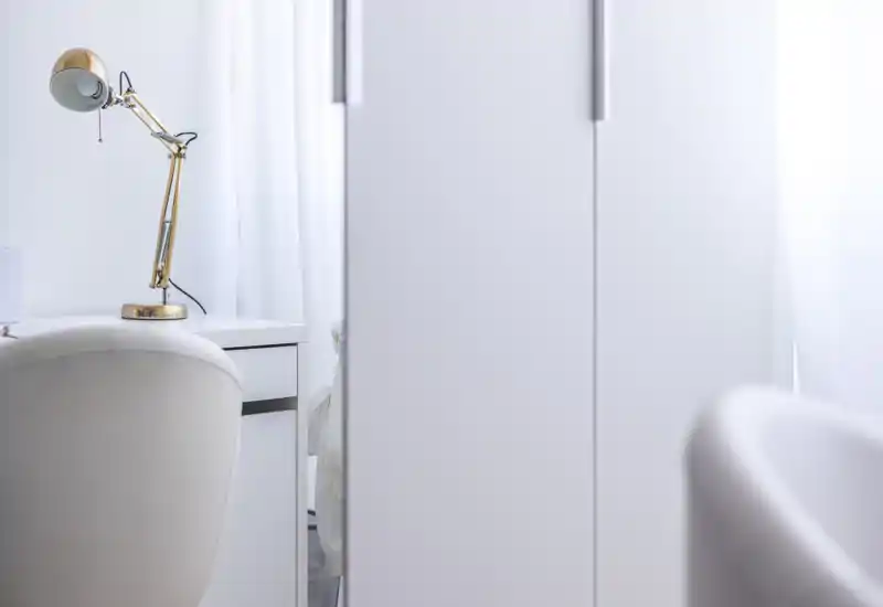 A bright, minimalist bedroom corner featuring a white desk, a gold desk lamp and soft seating; the bed is only partially visible through a narrow gap, giving a calm, airy feel.