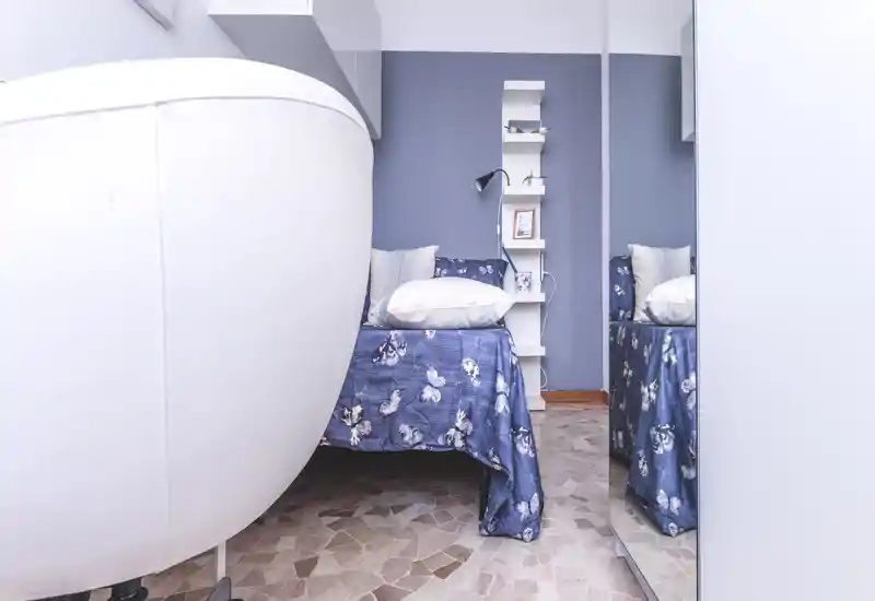 Compact bedroom with a single bed dressed in a blue floral bedcover, white pillows and a slim shelving unit against a soft blue wall; a large white chair is visible in the foreground and a mirrored wardrobe reflects the bed, creating depth.