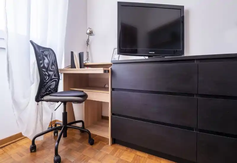 Compact bedroom corner with a black dresser, small wooden desk and swivel chair next to a bright window; simple, functional furnishings ideal for a single occupant or a student.
