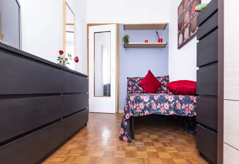 Bright, cozy bedroom with a floral-covered single bed and decorative red cushions, framed by dark wood dressers and parquet flooring.