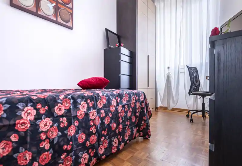 Cozy single bedroom with a floral bedspread, red accent pillow, wardrobe and a bright windowed corner with desk and chair — ideal for a student or solo guest.