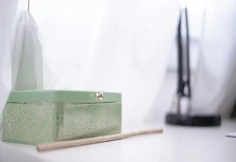 A close-up of a decorative green jewelry or storage box on a white surface with soft white curtains and a blurred lamp in the background. The image focuses on the object rather than a room layout.