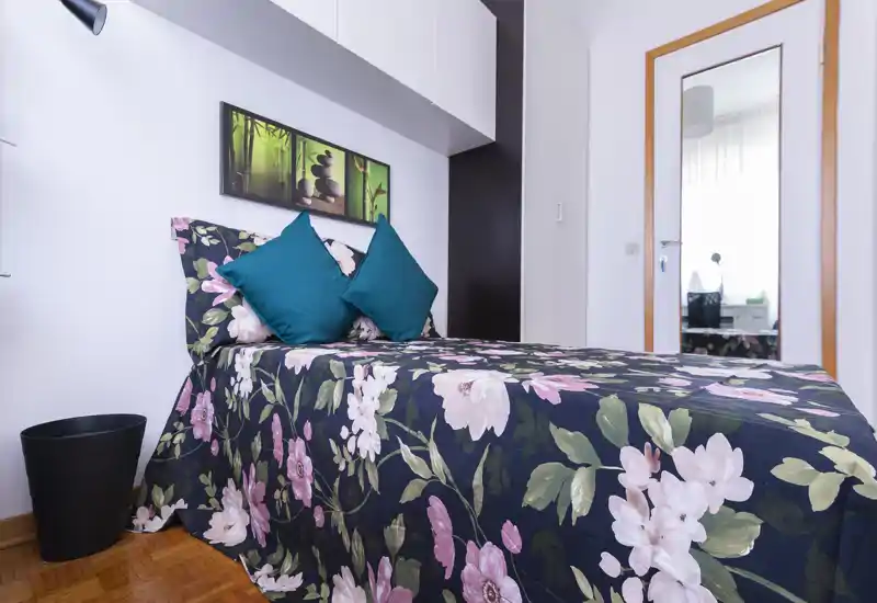 Bright, well-styled single bedroom with a floral bedspread, teal cushions and modern wall storage — ideal for a cozy one-person stay.