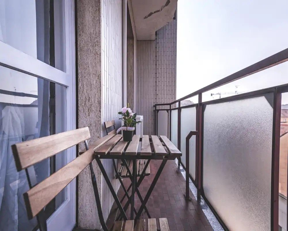 Balcón estrecho y luminoso con mesa bistró de madera y dos sillas, barandilla de vidrio esmerilado y maceta con flores — ideal para el café matutino.