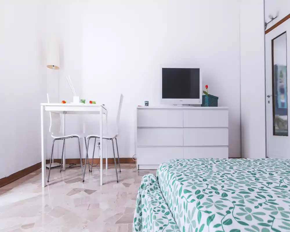 Dormitorio luminoso y minimalista con ropa de cama verde y blanca de estampado, cómoda baja con televisor y una pequeña mesa de trabajo/comedor — limpio y bien iluminado.
