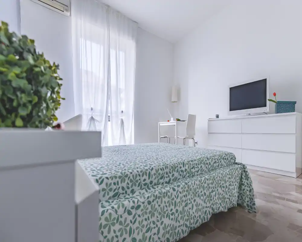 Dormitorio luminoso y minimalista con cama doble con funda estampada, cómoda con televisor y un pequeño escritorio junto a la ventana. Las cortinas translúcidas aportan mucha luz natural y una sensación tranquila y aireada.