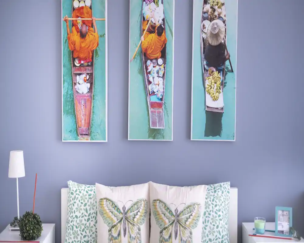 Rincón de dormitorio acogedor con cabecero y cojines decorativos con mariposas y cuadros verticales coloridos sobre la cama; iluminación suave y plantas junto a la mesita añaden calidez.