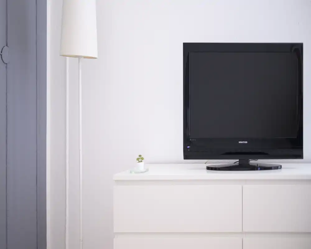 Zona giorno minimale con TV a schermo piatto su un comò bianco, lampada da terra e una piccola pianta decorativa — luminosa, pulita e moderna.