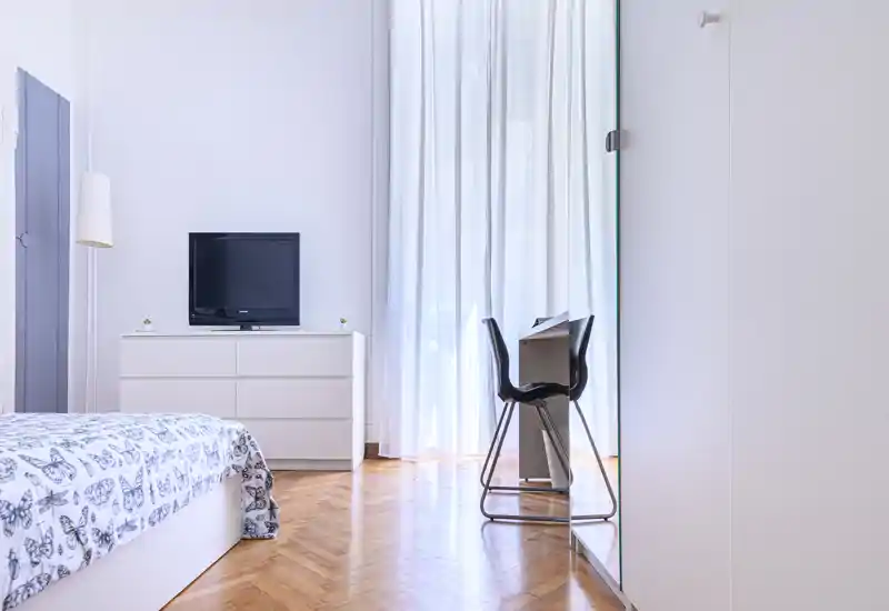 Bright, minimalist bedroom with patterned bedding, a dresser with TV, a small desk and chair by full-height sheer curtains — clean parquet flooring and soft natural light.