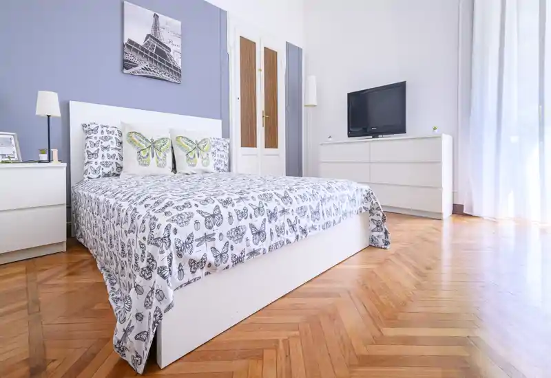 Bright, stylish bedroom with a large white bed dressed in butterfly-patterned linens, complementary nightstands and a dresser with TV. Hardwood floors and soft natural light create a warm, inviting atmosphere ideal for rental listings.