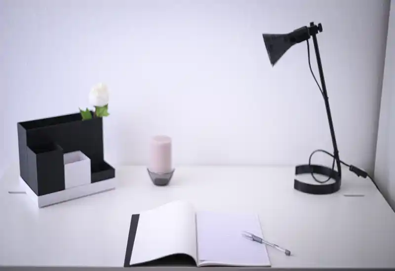 Minimal, bright workspace with a white desk, open notebook, pen, desk lamp and a small organizer with a candle — a calm, modern desk/working nook.