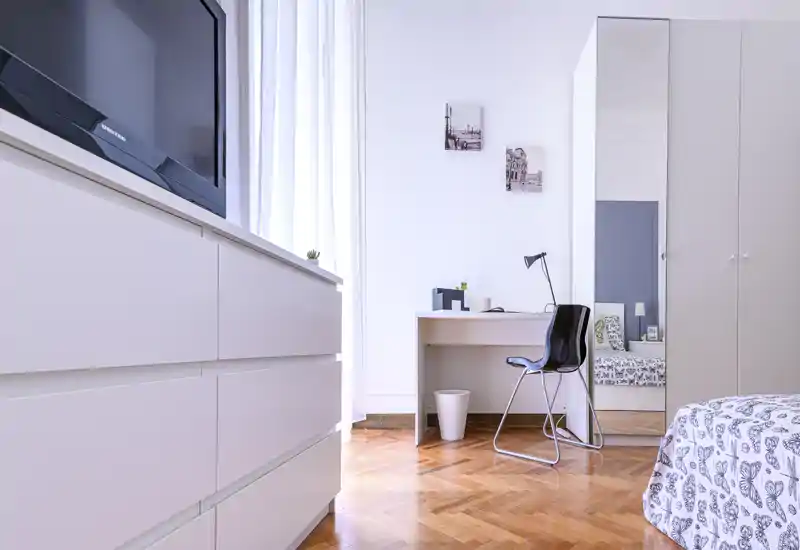 Bright, modern bedroom with patterned bedding, a desk and wardrobe with mirror; natural light highlights the parquet floor and minimalist furnishings.