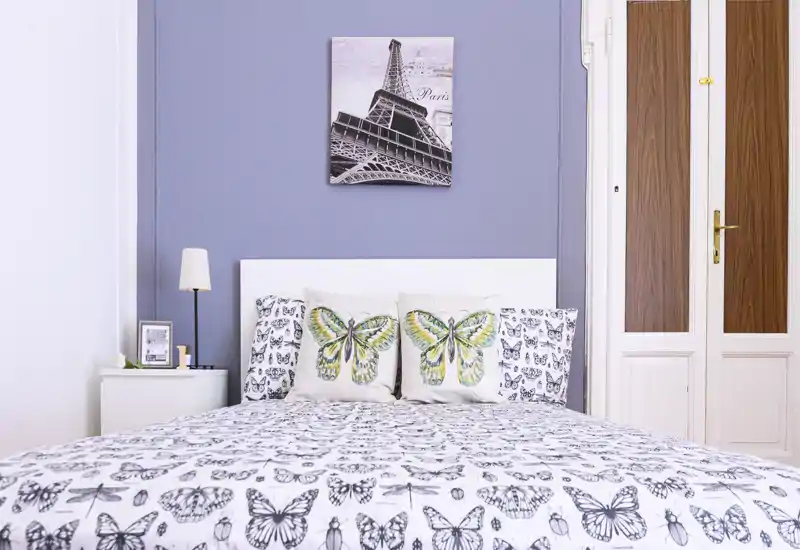 Bright, well-styled bedroom with a full bed covered in butterfly-patterned bedding, decorative cushions, a bedside lamp and a framed picture above the headboard.