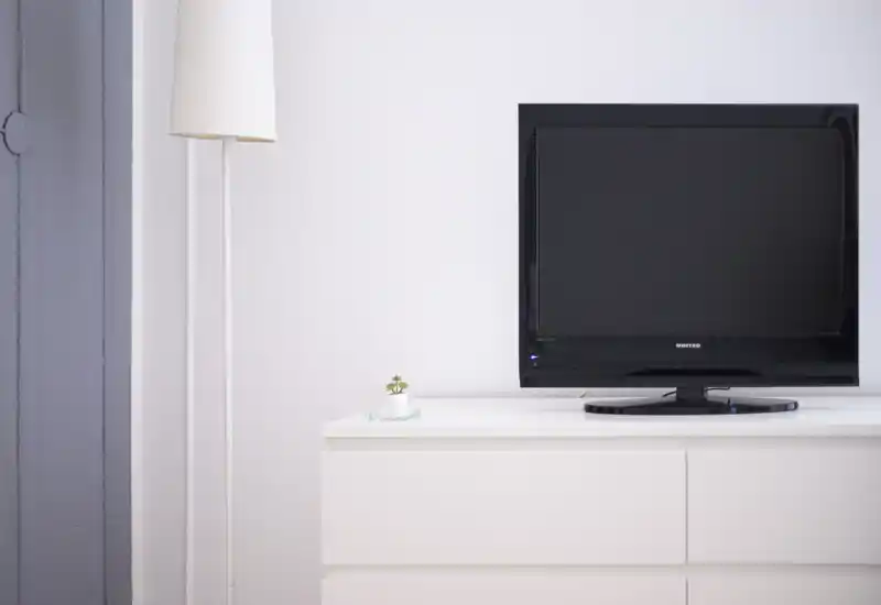 Minimal living area with a flat-screen TV on a white dresser, soft standing lamp and a small decorative plant — bright, clean and modern.