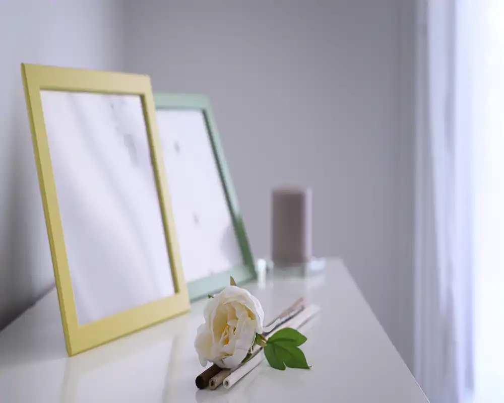 Close-up of decorative objects on a glossy surface: two empty picture frames, a candle and a white rose, with soft natural light from a nearby window.