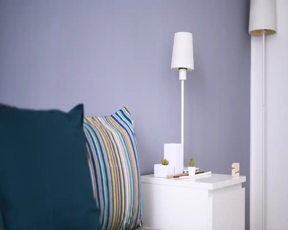 A calm bedroom corner with a striped accent pillow, bedside table and modern lamp — a cozy, well-lit spot ideal for relaxation.