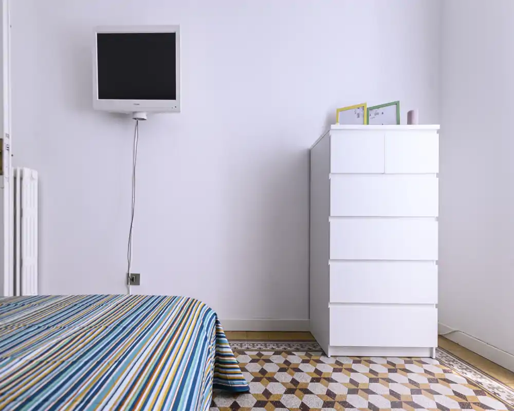 Bright, minimalist bedroom corner with a striped bed in the foreground, a wall-mounted TV and a tall white chest of drawers on a decorative tiled floor.
