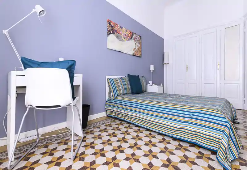 Bright, neatly styled single bedroom with striped bedding, decorative cushions and a small desk. The room features a patterned tiled floor and calming pastel wall for a modern, cozy feel.