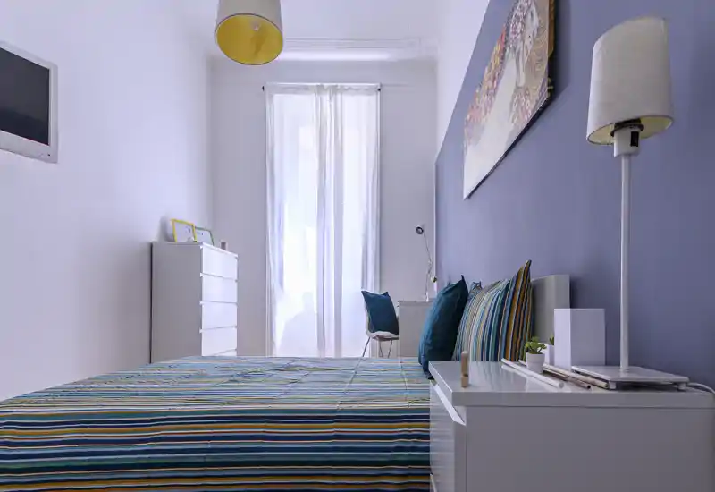 Bright, modern bedroom with a large striped bed in the foreground, coordinated cushions, bedside table and a tall window letting in soft natural light.