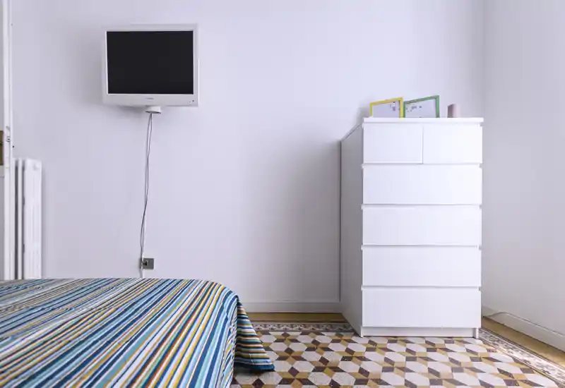 Bright, minimalist bedroom corner with a striped bed in the foreground, a wall-mounted TV and a tall white chest of drawers on a decorative tiled floor.