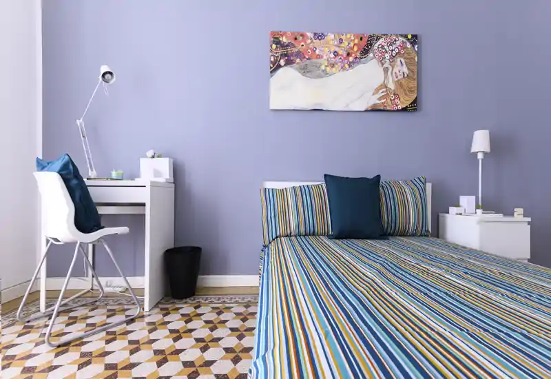 Bright, modern bedroom with a striped double bed, matching cushions and a small white desk. The room features soft lilac walls, decorative artwork above the bed and geometric patterned floor tiles.