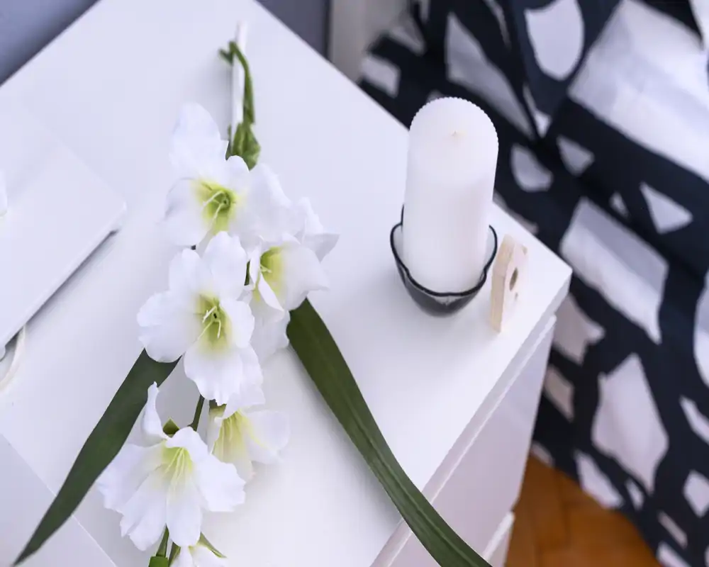 Angolo notte luminoso e curato con biancheria bianca, fiori decorativi e una candela su un comodino bianco — stile moderno e accogliente.