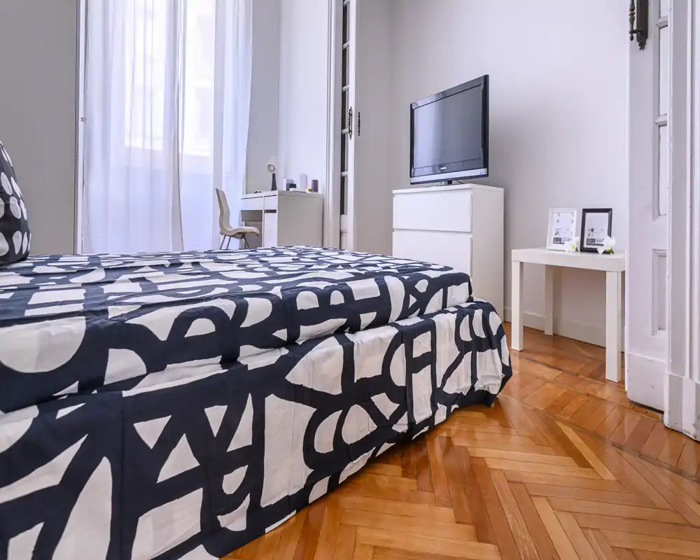 Camera da letto luminosa e moderna con letto matrimoniale a fantasia, pavimento in parquet a spina di pesce e TV su comò bianco. Ampia finestra con tende leggere e un piccolo scrittoio per un ambiente arioso.