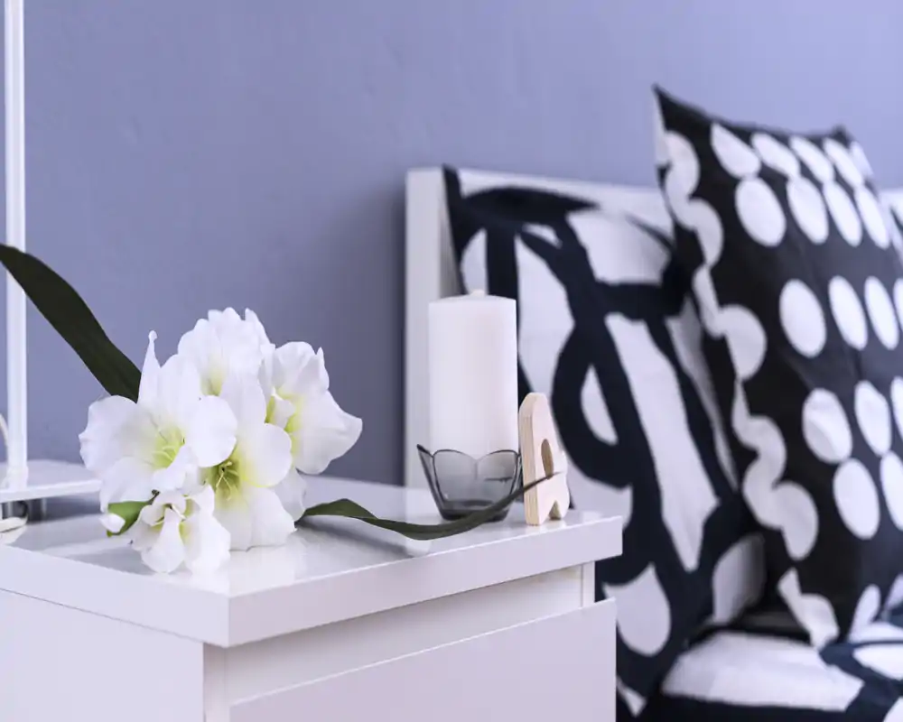 Primo piano del comodino di una camera da letto arredato: fiori bianchi freschi, una candela e cuscini con fantasia sul letto; luminoso, moderno e accogliente.