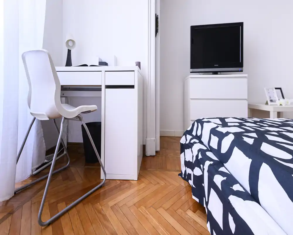 Camera da letto luminosa e moderna con biancheria a pattern, cassettiera con TV e una scrivania bianca compatta vicino alla finestra — perfetta per un affitto in città.