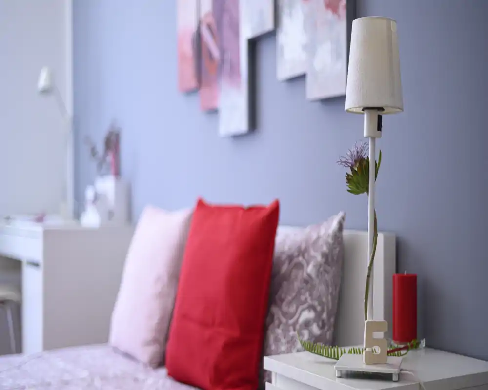 Bright, cozy bedroom close-up with patterned pillows and a red accent cushion on a neatly made bed, soft decorative lamp and wall art in the background.