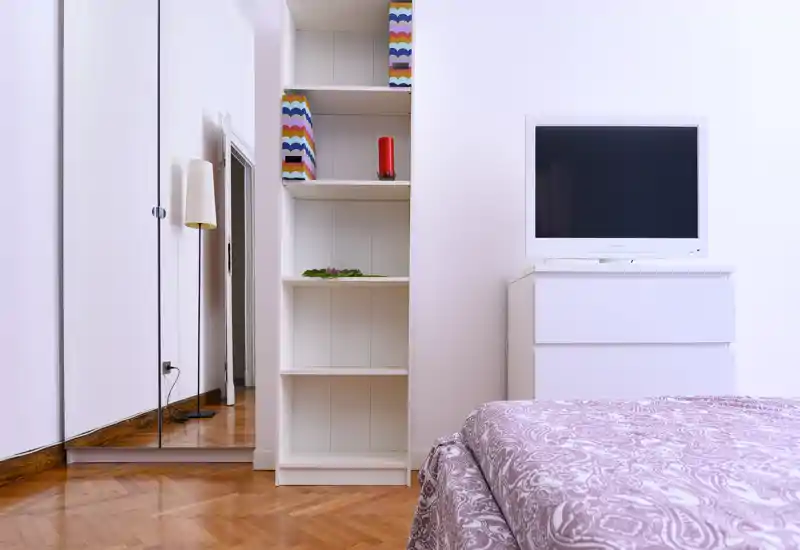 Bright, minimal bedroom corner with a patterned bedspread, a white chest with a TV and built-in shelving — a clean, modern setup ideal for a rental listing.