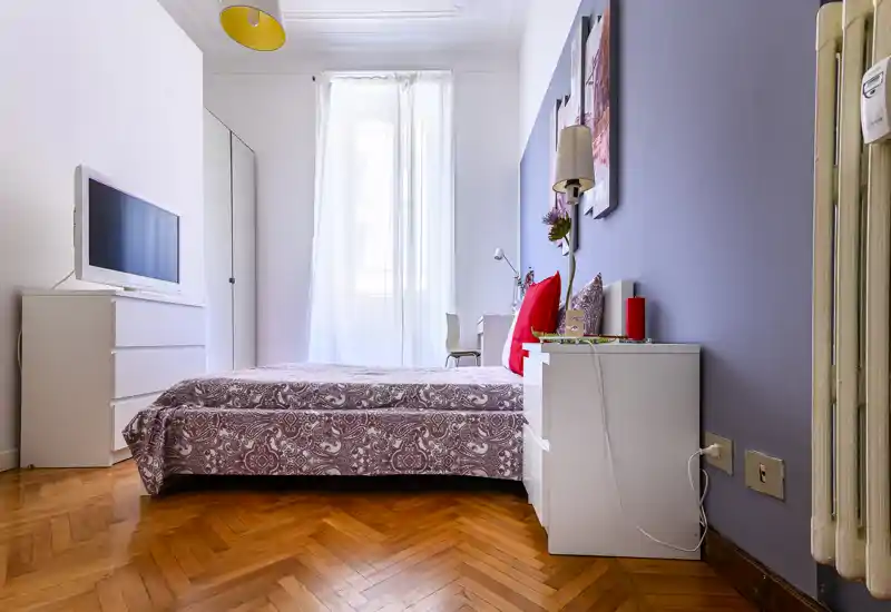 Bright, well-styled bedroom with a patterned double bed, bedside table and natural light from a tall window. The room features clean white furniture, warm parquet flooring and soft decorative cushions.